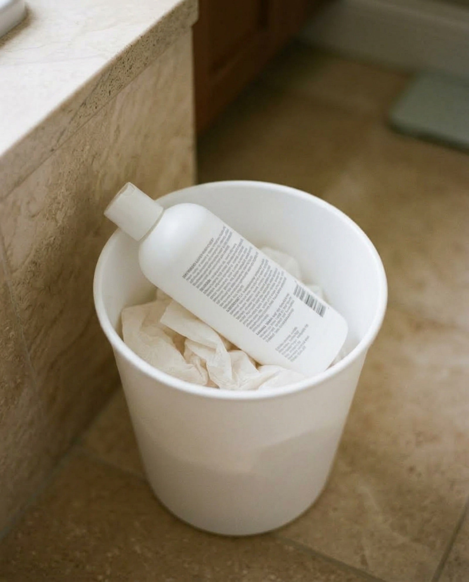 A discarded white sunscreen bottle lying in a small bathroom wastebasket on a tile floor